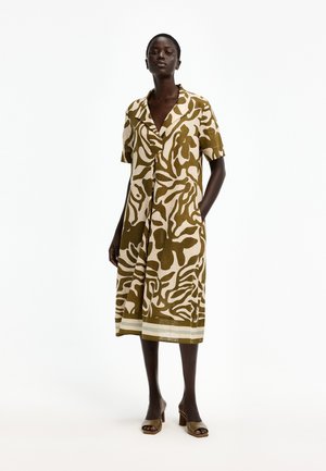 Frau steht in einem knielangen, kurzärmeligen Kleid mit grün-beigem abstraktem Blumenmuster, offenem Kragen und Taschen, kombiniert mit grünen Sandalen mit Blockabsatz.