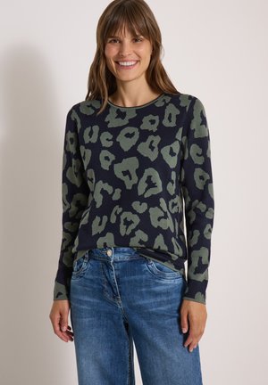 Een glimlachende vrouw met bruin haar draagt een donkere trui met lange mouwen met groene abstracte vlekken en een blauwe spijkerbroek tegen een effen achtergrond.