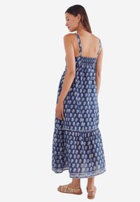Robe maxi en tissu bleu marine avec des motifs de feuilles blanches, bretelles réglables, dos smocké et un ourlet à volants en couches. Sandales plates dorées visibles.
