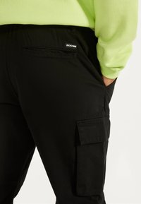 Mustat cargo-housut sivutaskuilla, valmistettu kestävästä kankaasta. Takataskussa on merkkilapuke, ja niitä on paritettu kirkkaanvihreän hupparin kanssa.