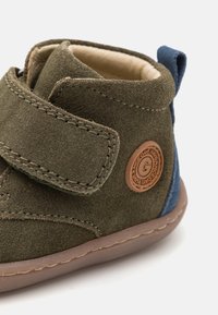 Bota Ankle verde em camurça com um detalhe azul no calcanhar, faixa em Velcro, sola de borracha bege e um emblema redondo de logo na lateral. Forro interior suave.