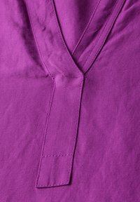 Fialová látka s hladkým povrchem, s výstřihem do V a prošívaným detailem, zdůrazňující jednoduchý design a čisté linie.
