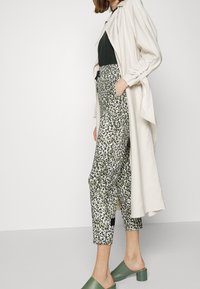 Beige lange jas, zwarte top, gemêleerde groen-witte broek en groene blokhak sandalen. De broek heeft een abstract bladerenontwerp.