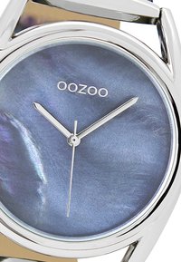 Montre bracelet ronde avec un boîtier en métal argenté, un cadran bleu nacré, des aiguilles argentées et le nom de la marque "OOZOO" sur le cadran.