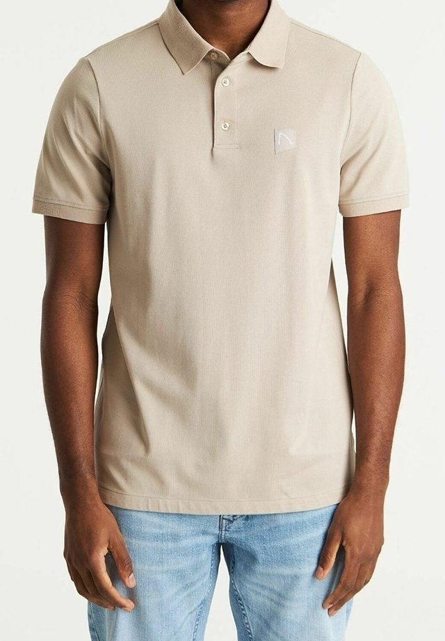 Homme portant un polo beige à manches courtes avec un col et des boutons, associé à un jean bleu clair, debout les bras détendus.