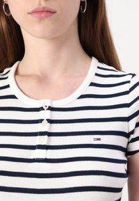 Triibuline lühikeste varrukatega topp valges ja tumesinises, ümar ribakrae ja viie nupuga, millel on väike logo vasakul.