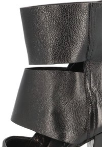 Black faux leather ankle boot featuring a high, split cuff design, a smooth texture, and a sleek silhouette with a metallic accent.