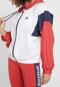 Vit och röd vindjacka med marina accenter. Har dragkedja framtill, elastiska manschetter och justerbar huvtröja. Reebok-logotyp på bröstet.