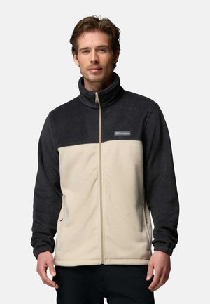 Mann trägt eine schwarze und beige Columbia Fleecejacke mit durchgehendem Frontreißverschluss und steht vor einem schlichten hellen Hintergrund.