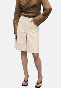 Shorts en coton beige avec une coupe droite, dotés de poches avant et d'une fermeture à bouton, associés à un top enveloppant marron et des sandales noires.