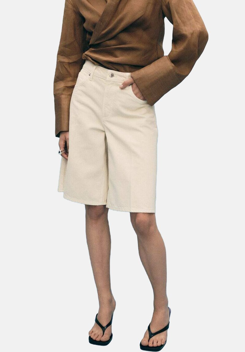 Shorts en coton beige avec une coupe droite, dotés de poches avant et d'une fermeture à bouton, associés à un top enveloppant marron et des sandales noires.