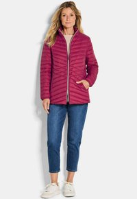 Femme portant une veste matelassée magenta, un jean cropped bleu et des baskets blanches, se tenant avec une main dans la poche de la veste.