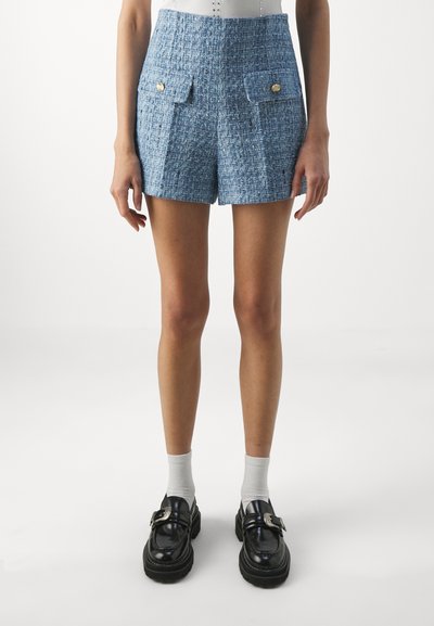 Shorts din tweed albastru cu un finisaj texturat, având două buzunare frontale și accente cu butoane aurii. Stilizate cu pantofi negri și șosete albe.