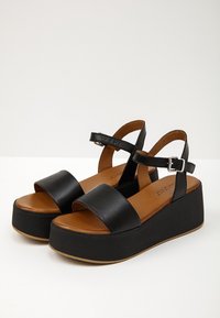 Černé platformové sandály s širokými předními pásky a pásky kolem kotníků, které mají stříbrné přezky, hnědé stélky a silné černé podrážky.