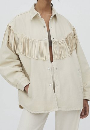 Persona que lleva una chaqueta beige suelta con botones y detalle de flecos largos en el pecho y los brazos, combinada con pantalones claros a juego.