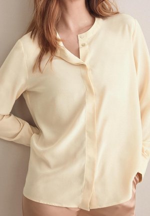 Donna che indossa una camicetta giallo chiaro con bottoni, maniche lunghe e scollo rotondo, mano appoggiata sull'anca.