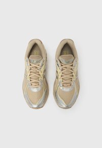 Ténis de corrida bege com parte superior em malha, detalhes prateados e biqueira arredondada. Apresentam atacadores e palmilhas acolchoadas para conforto. Sola de borracha.