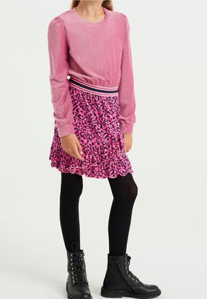 Un haut en velours côtelé rose à manches longues associé à une jupe rose à volants présentant un motif abstrait noir et rose, complété par des leggings et des bottes noires.