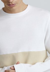 Sudadera blanca con una banda horizontal beige en la parte inferior. Mezcla de algodón, cuello redondo, puños acanalados y una textura suave.