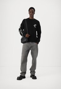 Pulover negru cu motive grafice albe pe mâneci, purtat cu blugi gri lejeri și pantofi masivi negri. Geantă de umăr minimalistă, neagră.