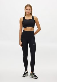 Svart sport-BH med breda band och logotyp, kombinerad med högmidjade svarta leggings med fickor. Modell bär träningsskor.