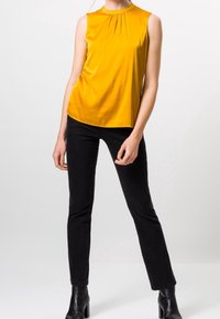 Gelbes ärmelloses Top mit gerafftem Ausschnitt, kombiniert mit schwarzen, enganliegenden Jeans und Stiefeletten. Glattes, glänzendes Material mit einer eleganten Silhouette.