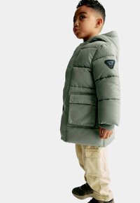 Barn i grøn polstret vinterjakke med hætte, beige cargo bukser og sorte sneakers, der står sidelæns på hvid baggrund.