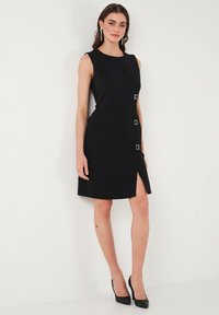Black sleeveless dress with a fitted silhouette, featuring square hardware accents and a side slit. Smooth fabric with a sleek finish.