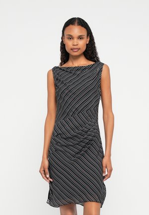 Hosszú, göndör hajú nő, akit ujjatlan fekete ruhában láthatunk, amelyen átlós fehér és kék csíkok vannak, sima háttér előtt.