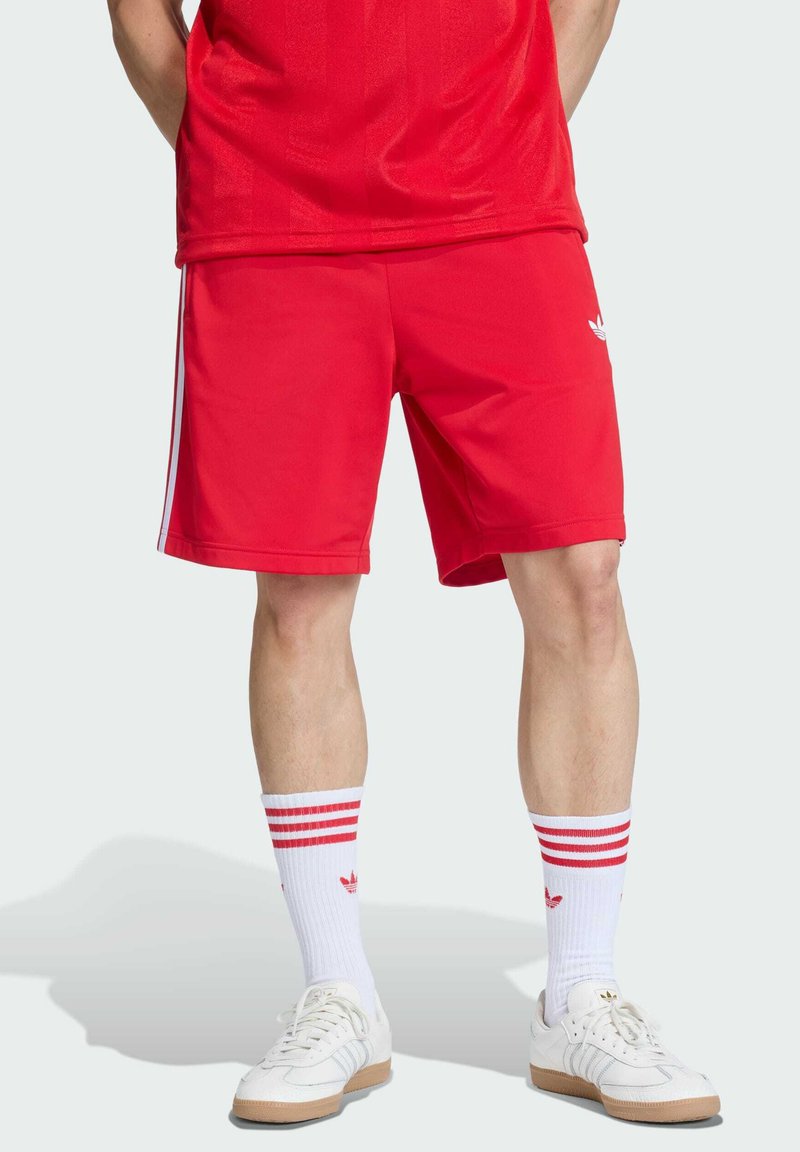 Shorts de sport rouges avec des bandes latérales, portés avec des chaussettes blanches ornées de rayures rouges et d'un logo, assortis à des baskets blanches.