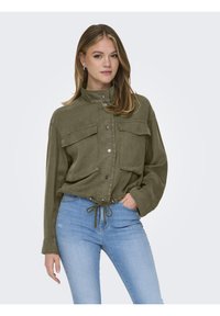 Giacca verde oliva, con bottoni e colletto alto, due tasche frontali, vita regolabile con cordino e taglio morbido, abbinata a jeans azzurri chiari.