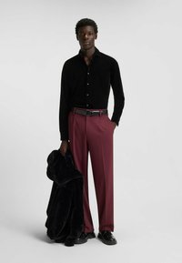 Schwarzes Cordhemd, bordeauxrote Hose, schwarzer Gürtel, hält eine schwarze Kunstpelzjacke. Glatte Texturen und kontrastierende Farben.