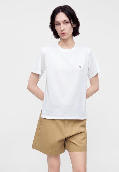 Personne aux cheveux courts et foncés portant un t-shirt blanc uni avec un petit logo noir et un short beige, debout les mains derrière le dos.