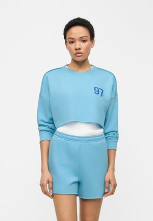 Kvinde iført lyseblå cropped sweatshirt med "97" tryk og matchende shorts, stående mod en ensfarvet hvid baggrund.