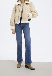 Beżowa pikowana kurtka bomberka z białym sztucznym futerkiem, czarnymi dodatkami, w połączeniu z niebieskimi rozszerzanymi jeansami i zielonymi botkami za kostkę.