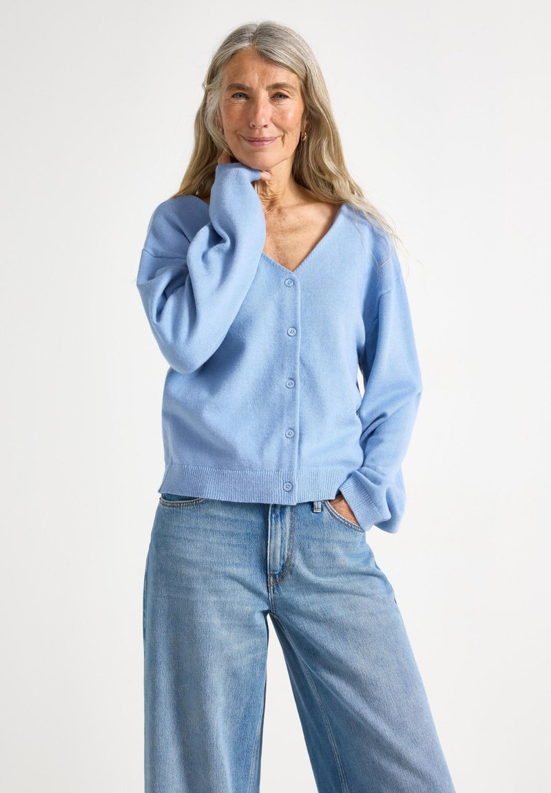 Cardigan bleu clair avec décolleté en v, devant boutonné, manches longues et ourlet côtelé. Associé à un jean bleu ample.