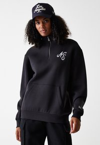 Schwarzer hoodie mit Reißverschluss, gerippten Bündchen und Saum, mit weißem gesticktem Logo auf der Brust und einer vorne angebrachten Kängurutasche.