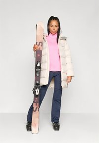 Løs, hvit jakke, rosa høyhalset genser, marineblå skibukser, svarte skistøvler. Holder ski med svarte bindinger og rosa detaljer.