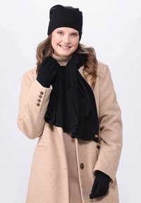 Beanie lavorato a maglia nera, sciarpa e guanti abbinati a un cappotto beige; tessuti texture; aspetto liscio e caldo; la sciarpa si adagia intorno al collo.