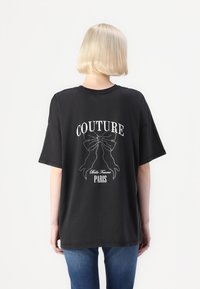 Juoda per didelį T-marškiniai su balta užrašu "COUTURE" ir kaspino dizainu, papuoštu rhinestones; švelni medvilnė ir trumpi rankovės.