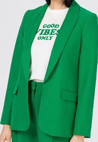 Zelený blejzr s přiléhavým střihem, s klopami a bočními kapsami, v kombinaci s bílým tričkem, na kterém je zelený nápis: "POUZE DOBRÉ VNEMENÍ."
