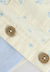 Material crem cu un model floral albastru, dotat cu nasturi bej. Marginea de material albastru deschis este vizibilă dedesubt, având texturi și cusături contrastante.