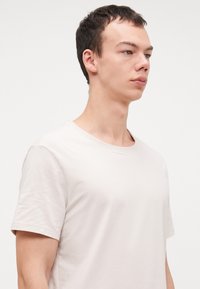 Helles beige Baumwoll-T-Shirt mit Rundhalsausschnitt und kurzen Ärmeln, das eine glatte Textur und ein schlichtes, klassisches Design aufweist.