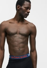 Μυώδης άνδρας με σκούρο δέρμα φορώντας μαύρα εσώρουχα Björn Borg, κοιτάζοντας προς τα κάτω και αριστερά σε απλό γκρι φόντο.