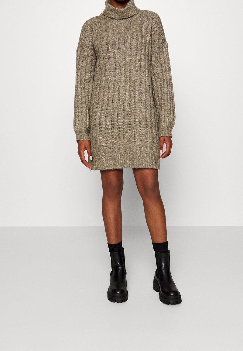 Femme portant une robe pull à col roulé beige côtelée à manches longues, associée à des bottines noires épaisses et des chaussettes noires.