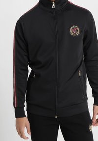 Black zip-up jacket made of synthetic material, featuring red stripes on sleeves, gold zippers, and a logo patch on the chest.