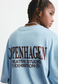 Hellblaue Baumwoll-T-Shirt mit oversized Passform, auf der Rückseite mit auffälligem braunem Text: "COPENHAGEN CREATIVE STUDIO EXHIBITION".