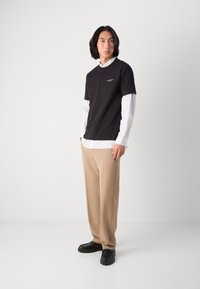 Black short-sleeve t-shirt over a white striped long-sleeve shirt, paired with beige trousers and black shoes. Simple design with minimal accents.