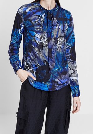 Femme portant un chemisier bleu à manches longues à motif floral avec un col noué et un pantalon cargo noir, main dans la poche.