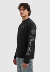 Zwart shirt met lange mouwen met een ronde hals, witte bedrukte tekst langs de rechter mouw en een klein geborduurd logo op de borst.